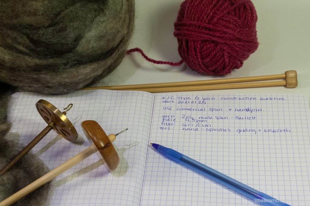 a ball of fiber, a ball of yarn, knitting needles, two spindles on top of open notebook