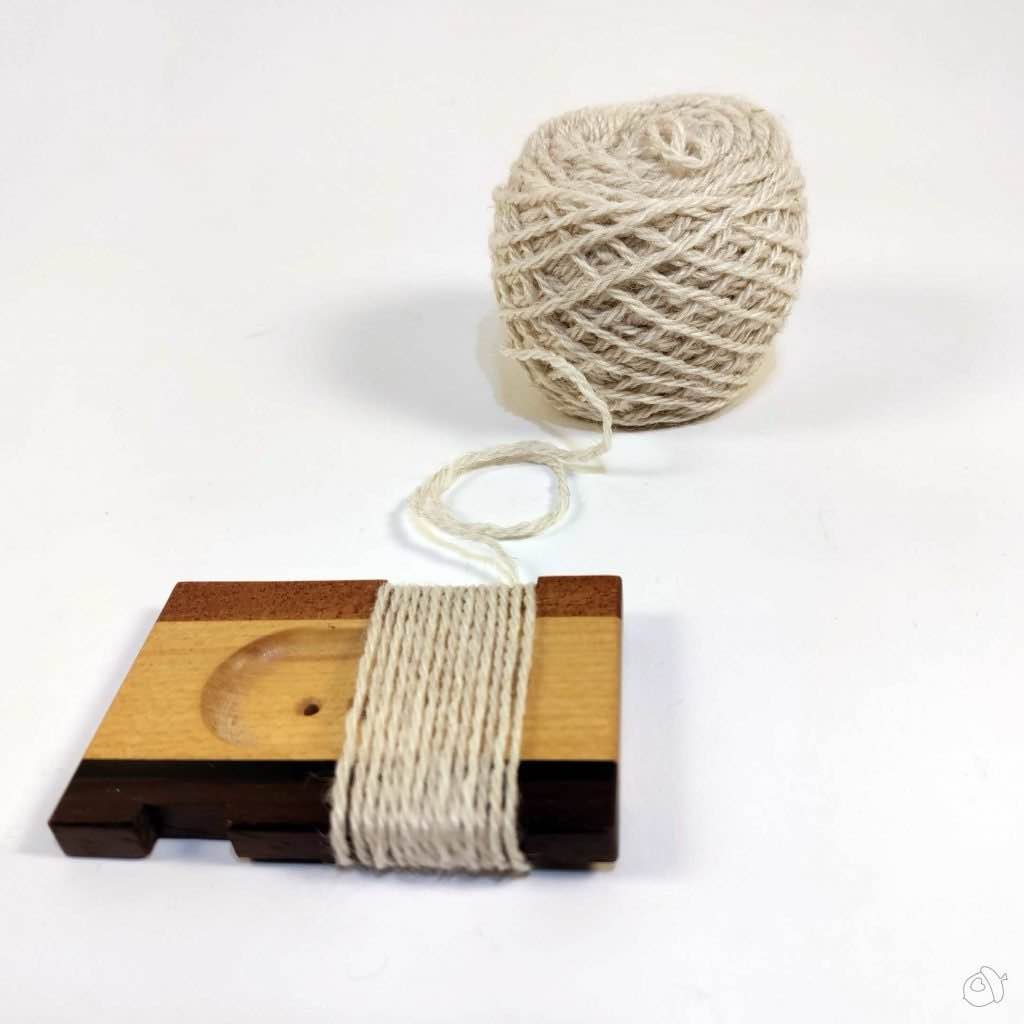 Leftover cake of LGF Suri Sock in neutral white and WPI tool with yarn wrapped around it on a plain white background