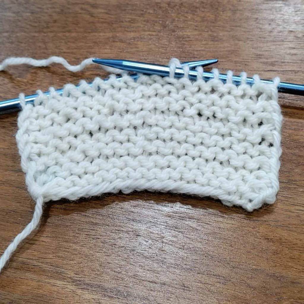 A white yarn being knit into a garter stitch swatch. The knitting needles are a light blue aluminum. The swatch is on a wooden table.