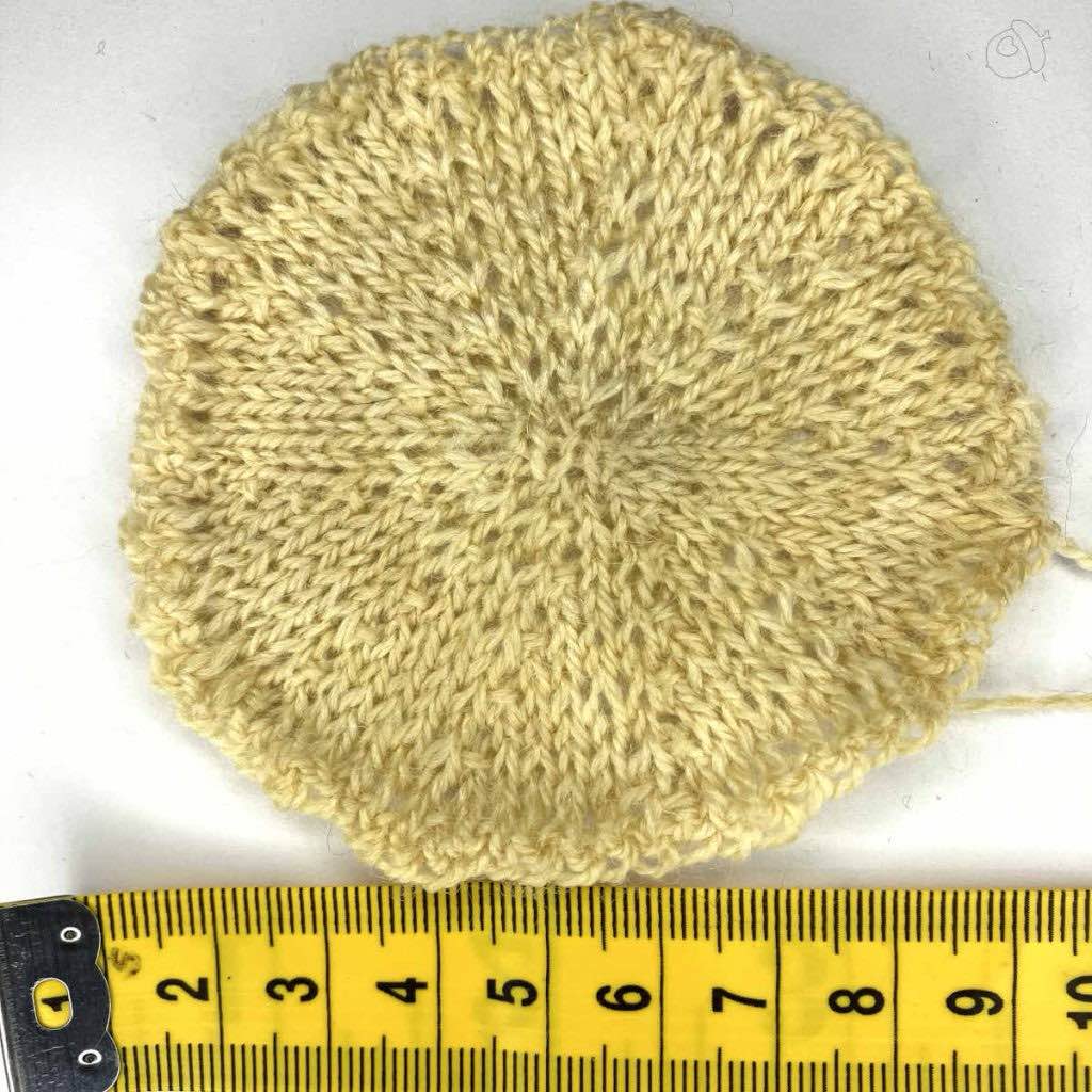stockinette knit circular swatch. the yarn color is off-white the background is white. there is a yellow tape ruler showing 10 cm. 