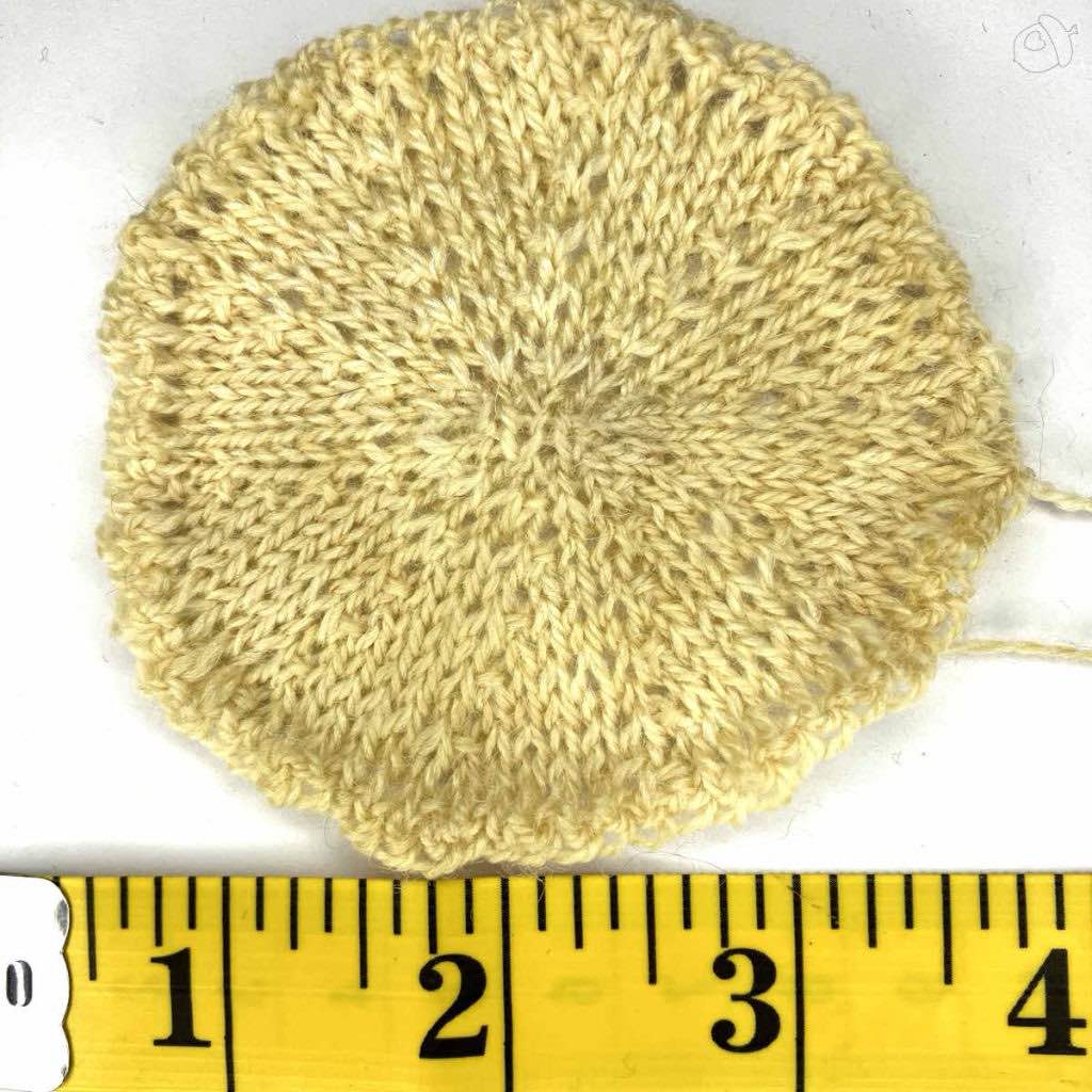 stockinette knit circular swatch. the yarn color is off-white the background is white. there is a yellow tape ruler showing 4 inches. 